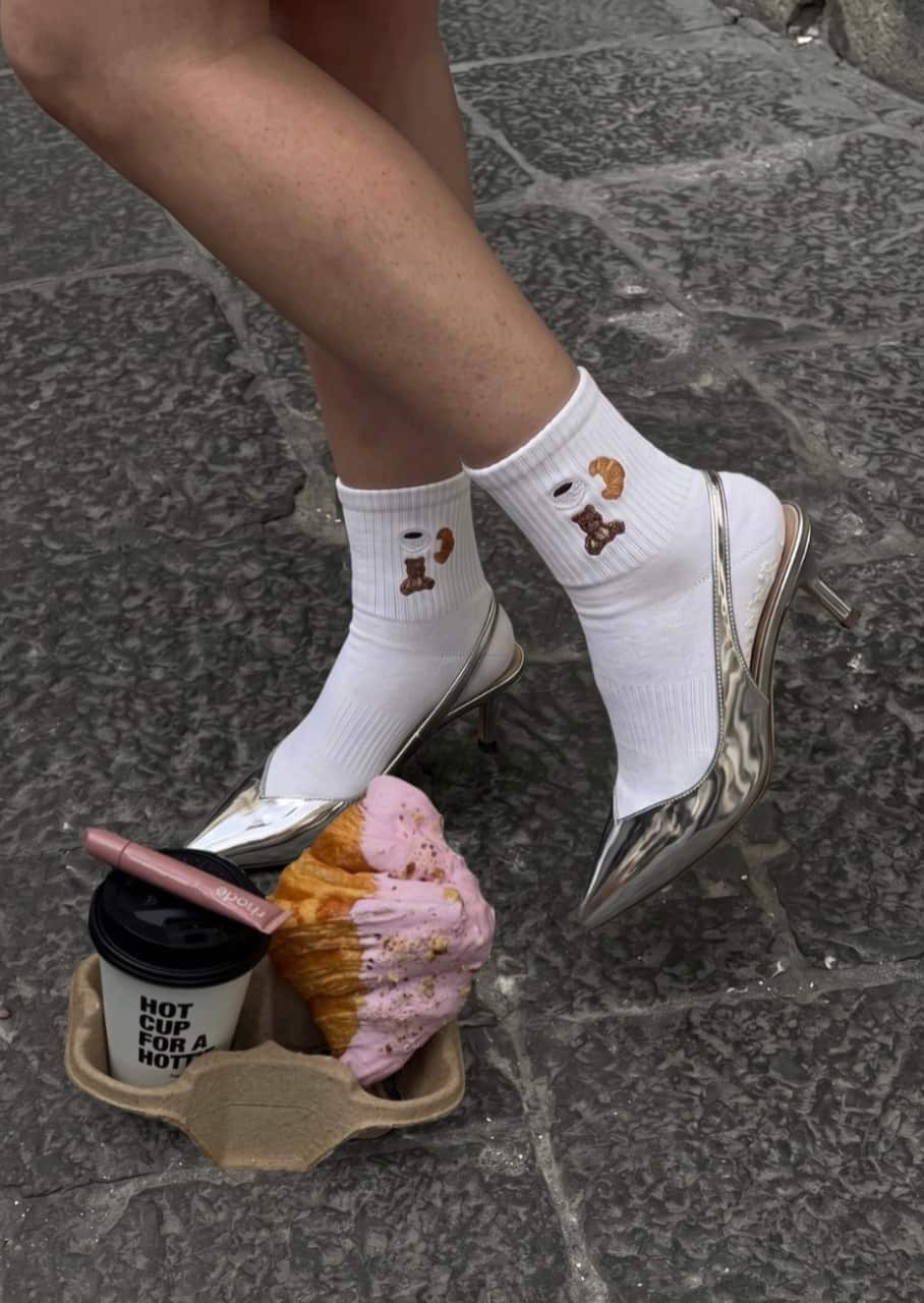 Weiße Grippy Socken mit Bärchen-Motiv kombiniert mit silbernen High-Heels, Kaffee und einem Croissant auf einem Steinboden