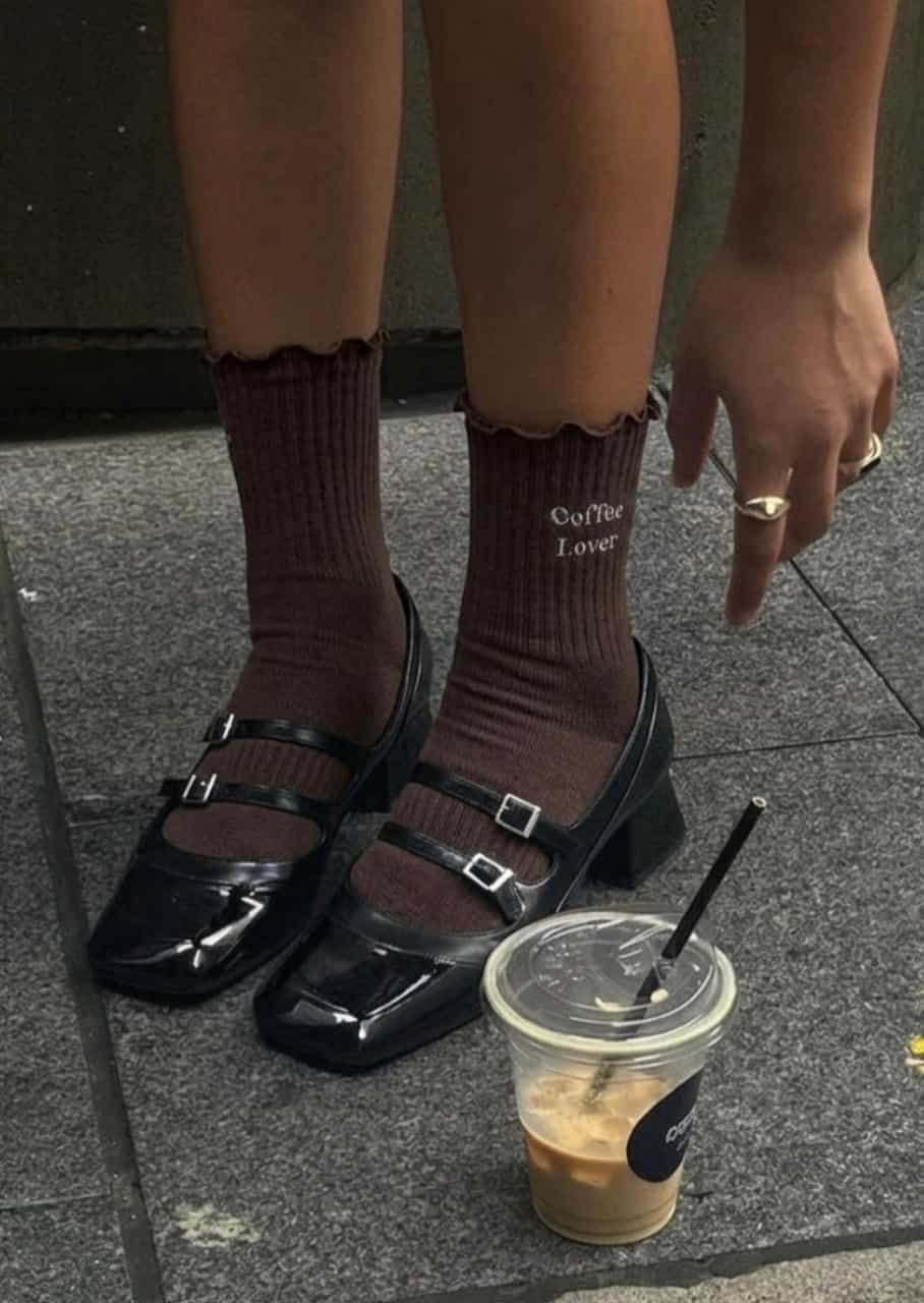Braune Grippy Socken mit „Coffee Lover“-Schriftzug in schwarzen Mary-Jane-Schuhen neben einem Eiskaffee auf einem Steinboden.