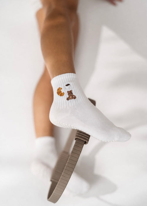 Weiße Pilates Socken mit einem Teddybären, Croissant und Kaffee. Zwischen den Knöcheln befindet sich ein Pilates Ring