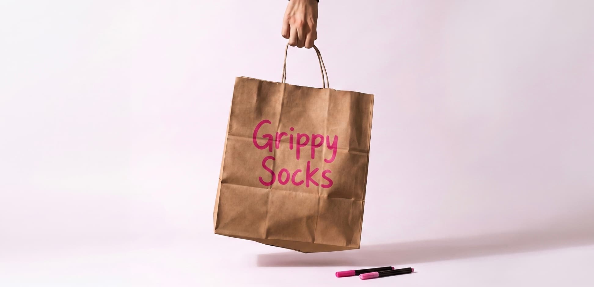 Braune Papiertüte mit pinkem Schriftzug „Grippy Socks“, die von einer Hand vor einem hellen Hintergrund gehalten wird.