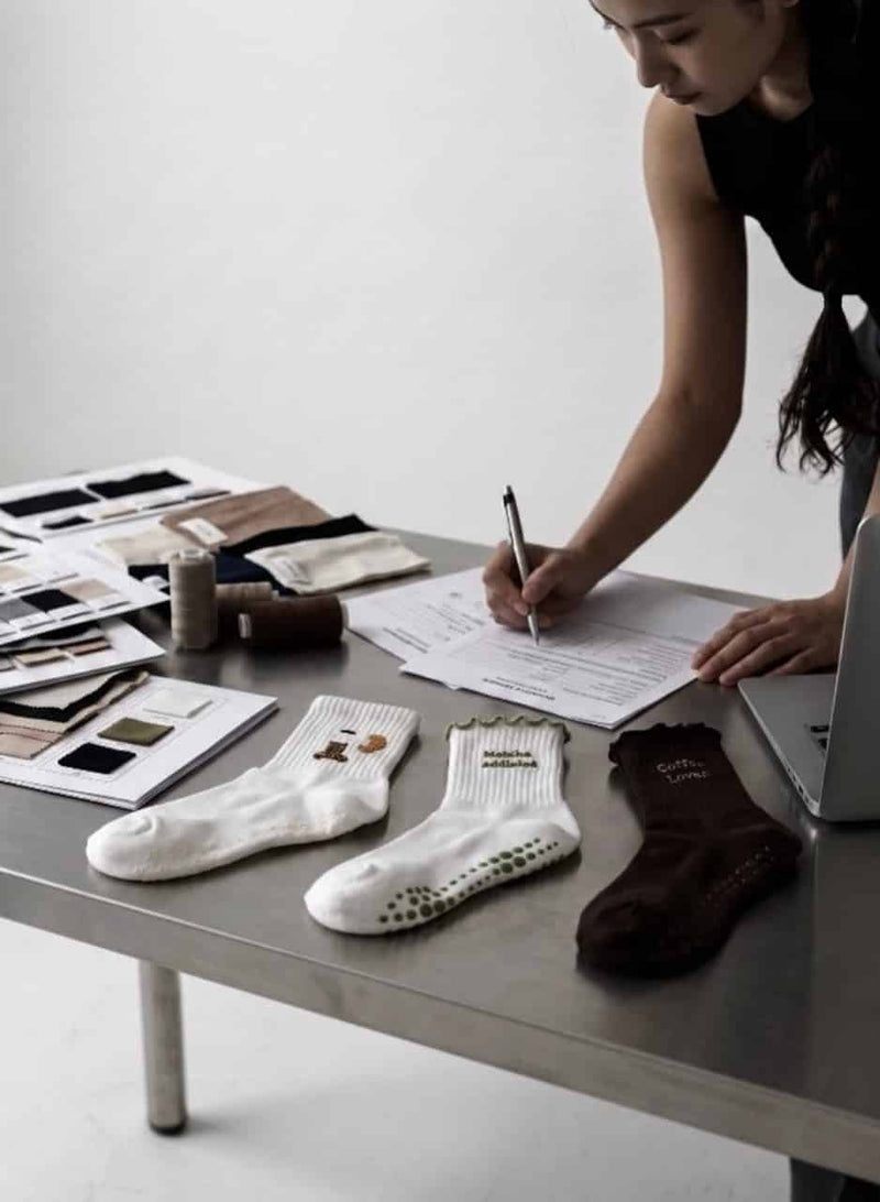 Person arbeitet an einem Tisch und dokumentiert die Entwicklung von Pilates-Grip-Socken. Auf dem Tisch liegen verschiedene Stoffmuster, Garnrollen, Unterlagen, ein Laptop sowie drei Socken in Weiß und Braun mit unterschiedlichen Designs und rutschfesten Noppen.