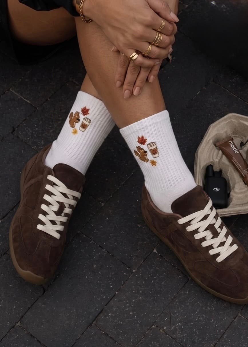 Weiße gerippte Socken mit herbstlichem Stickmotiv aus Kaffeebecher, Eichhörnchen und bunten Blättern, kombiniert mit braunen Sneakern und goldenen Schmuckdetails.