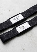 Schwarze Zughilfen von NST Fitness mit grauem Wabenmuster und weißem Logo-Label für Krafttraining und Gewichtheben.