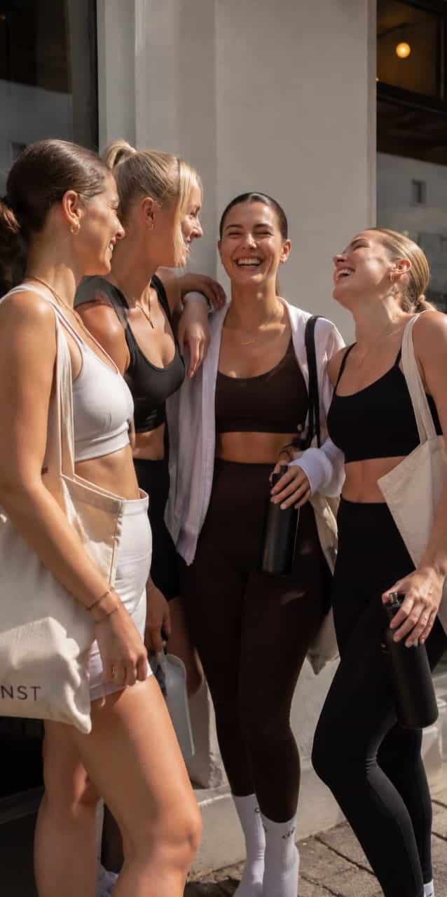 Vier Frauen in sportlicher Kleidung stehen draußen zusammen, lachen und unterhalten sich. Sie tragen schwarze und weiße Outfits, einige halten Trinkflaschen in der Hand und tragen Stofftaschen.