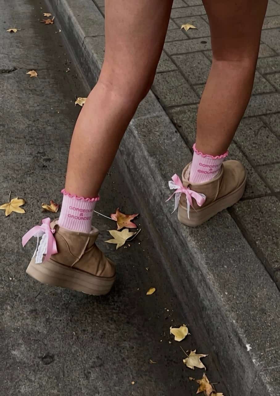 Rosa Grippy Socken mit gewelltem Rand und „GORGEOUS & GROUNDED“-Schriftzug, kombiniert mit beigen Plateau-Schuhen mit rosa Schleifen auf einer Straße mit Herbstblättern.