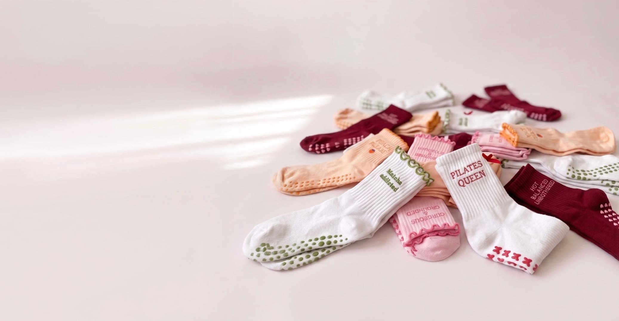 Verschiedene farbenfrohe Grip-Socken von NST liegen auf einer hellen Oberfläche, darunter weiße, rosa, burgunderrote und orangefarbene Pilates-Socken mit unterschiedlichen Designs und Anti-Rutsch-Noppen.