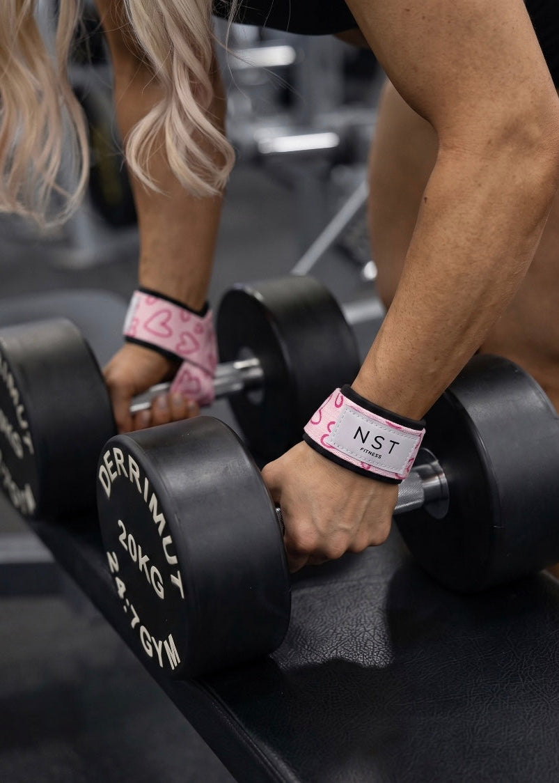 Frau trainiert mit schwarzen Kurzhanteln im Gym und trägt rosa NAST Fitness Zughilfen mit pinkem Herzmuster an beiden Handgelenken.