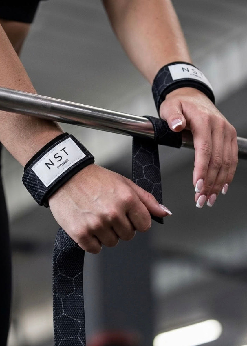 Frau verwendet schwarze NST Fitness Zughilfen mit grauem Muster beim Krafttraining an einer Langhantel. Die gepolsterten Zughilfen unterstützen den Griff und bieten zusätzlichen Halt beim Gewichtheben.