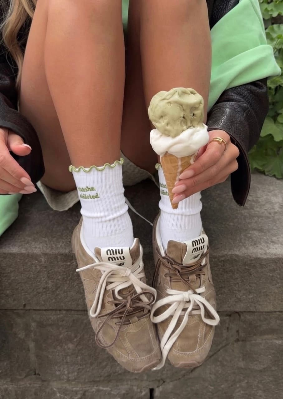 Weiße Grippy Socken mit grünem „Matcha Addicted“-Schriftzug und gewelltem Rand, kombiniert mit beigefarbenen Sneakern und einem Matcha-Eis in der Hand.