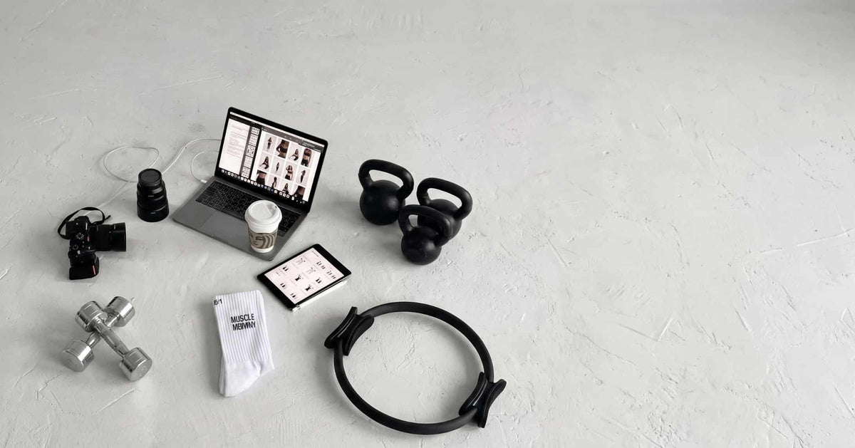 Weiße Sportsocken mit „Muscle Mommy“-Schriftzug neben Kurzhanteln, Kettlebells, Pilates-Ring, Kamera und Laptop in einem Fitness-Content-Setup
