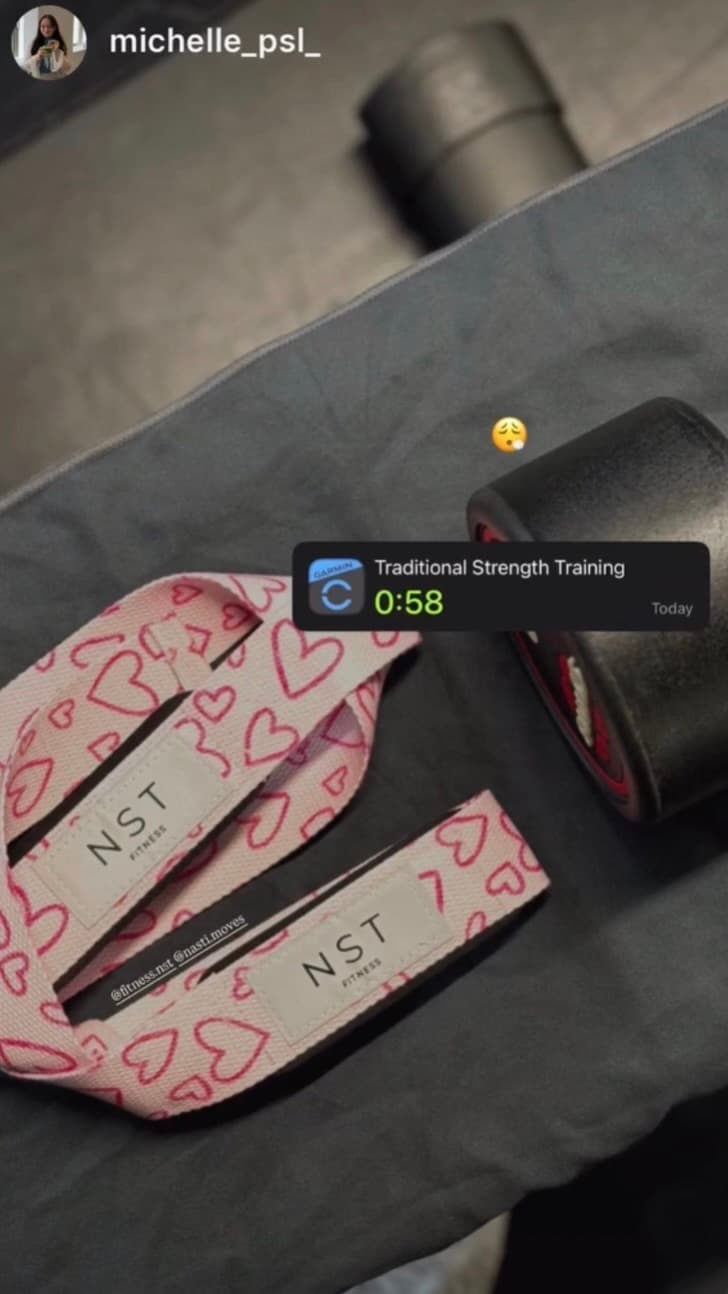 Rosa NST Fitness Zughilfen mit pinkem Herzmuster und Logo neben einer Hantel auf einer Trainingsbank – stylisches Gym-Zubehör für Frauen beim Krafttraining.