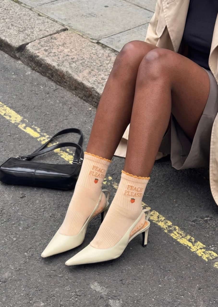 Pfirsichfarbene Grippy Socken mit „PEACH PLEASE“-Schriftzug und kleinem Pfirsichmotiv, kombiniert mit cremefarbenen High Heels und einem schwarzen Handtaschen auf der Straße.
