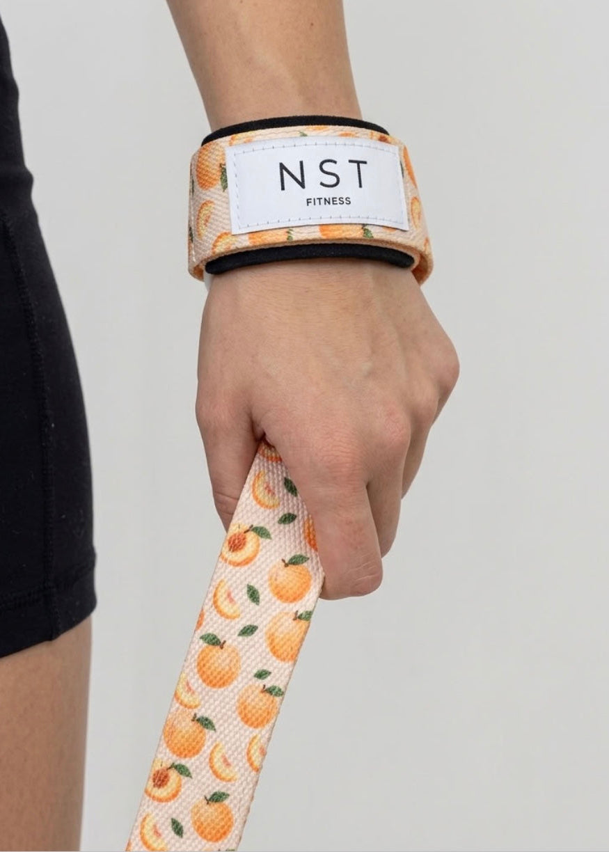 Detailaufnahme einer Hand mit NST Fitness Zughilfe im verspielten Pfirsich-Print, die beim Krafttraining für zusätzlichen Halt am Handgelenk sorgt.