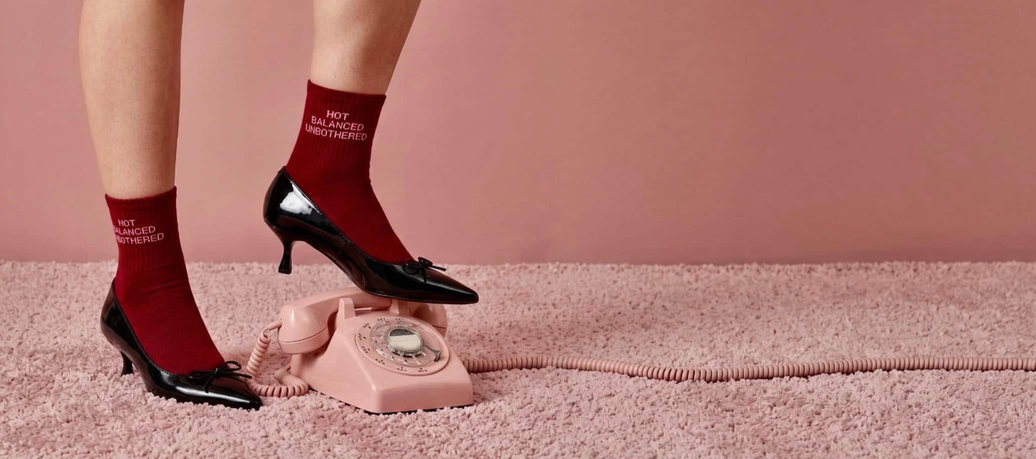 Rote Grip-Socken mit „Hot Balanced Unbothered“-Schriftzug kombiniert mit schwarzen High Heels auf einem rosa Teppich neben einem nostalgischen rosa Telefon.