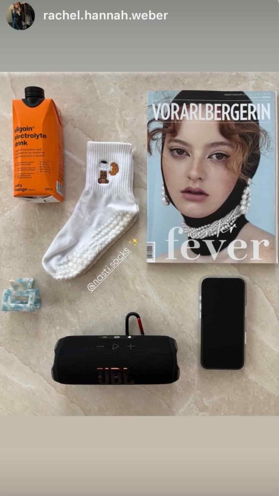 Weiße NST Grip Socks neben einem Magazin, einem Elektrolytgetränk, einem Smartphone und einem tragbaren Lautsprecher auf einem hellen Tisch.