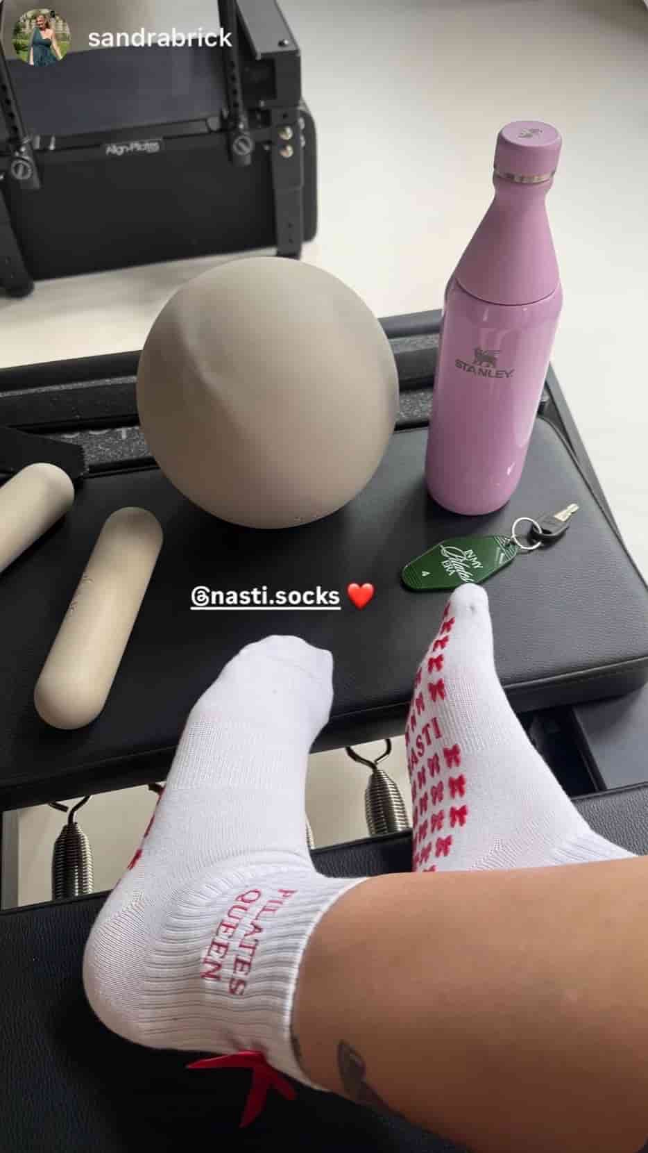 Weiße Pilates-Socken mit rotem Schleifen- und Schriftzug-Design an den Füßen auf einem Reformer. Im Hintergrund sind ein Pilates-Ball, eine Trinkflasche und weiteres Pilates-Zubehör zu sehen.