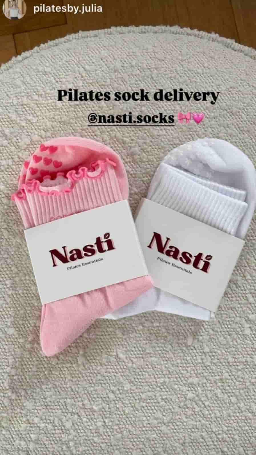 Zwei verpackte NST Pilates-Socken in Rosa und Weiß liegen auf einem Teppich. Die Socken haben dekorative Designs und rutschfeste Sohlen; darüber steht „Pilates sock delivery“