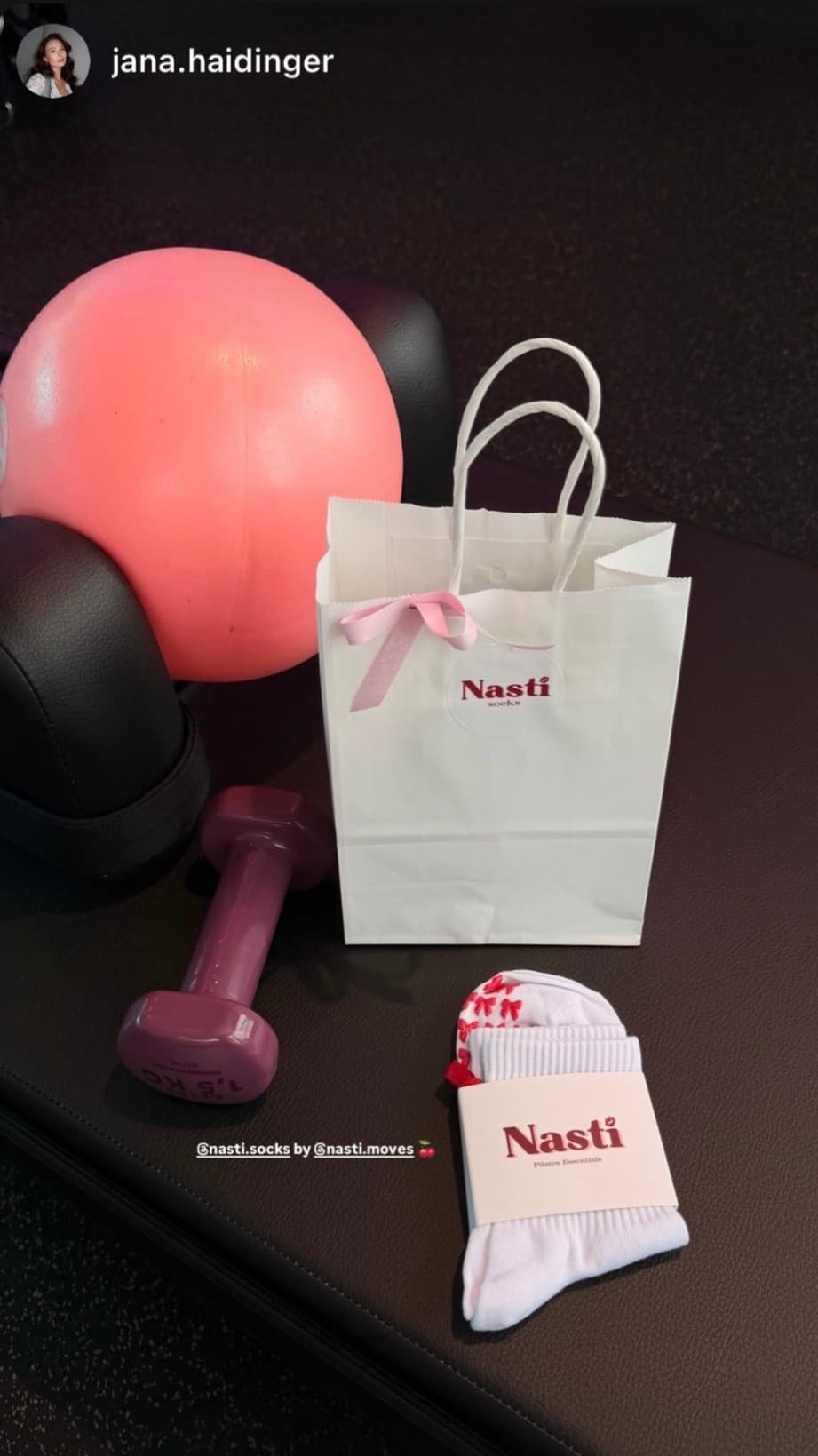 NST Pilates Grip Socken in weißer Verpackung mit pinkem Blumenmuster neben einer NST Geschenktüte und Fitness-Equipment.