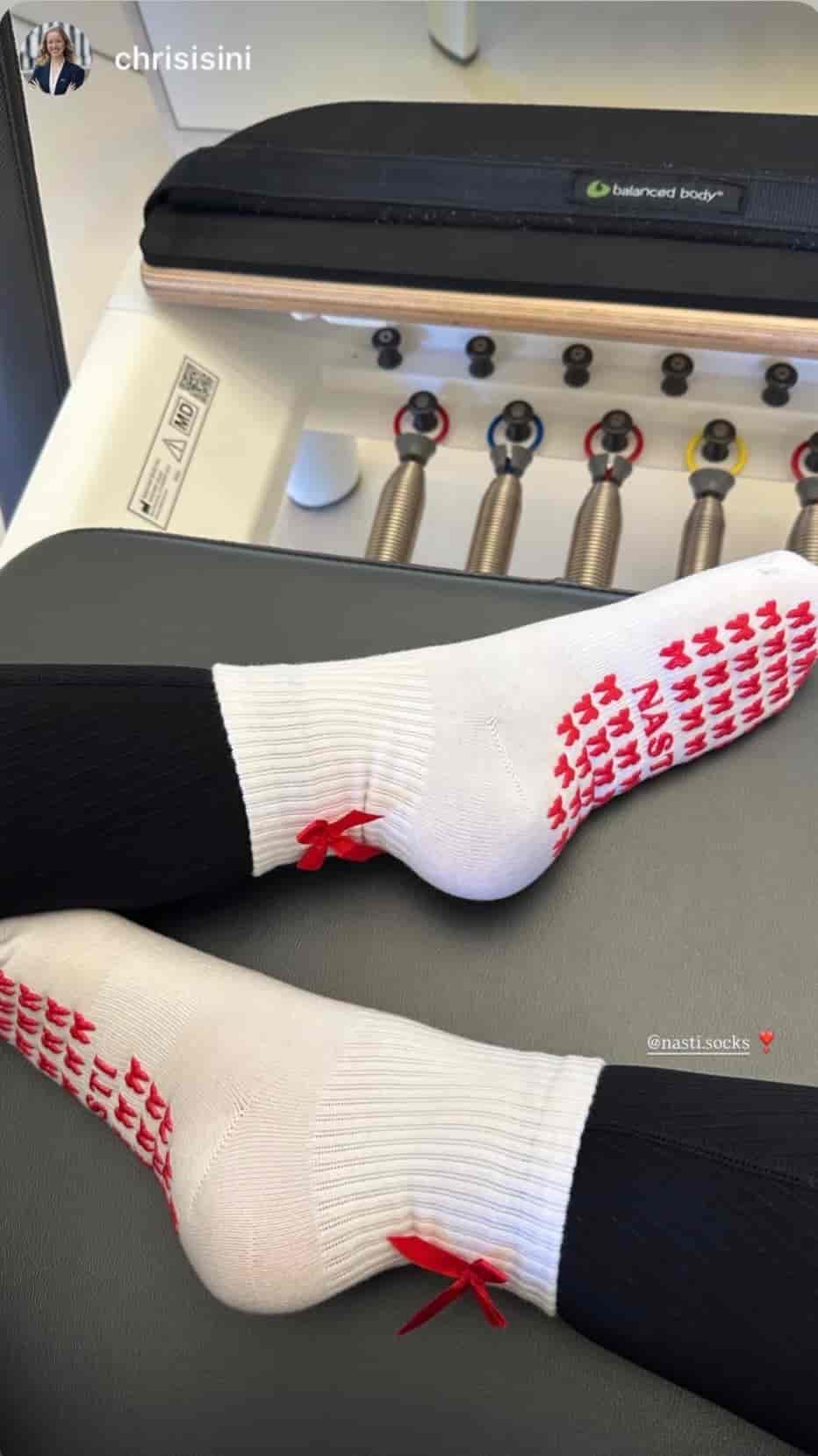 Zwei Füße in weißen Pilates Grip Socks mit roten Schleifen und rutschfestem Grip auf einem Reformer im Pilates-Studio.