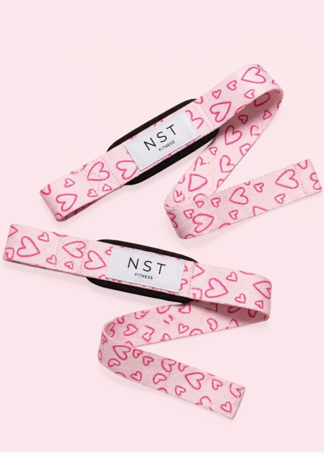 Rosa NST Fitness Zughilfen mit pinkem Herzmuster und weißem Logo, dekorativ auf hellrosa Hintergrund arrangiert. Stylische Lifting Straps für Krafttraining, Gym und Gewichtheben.
