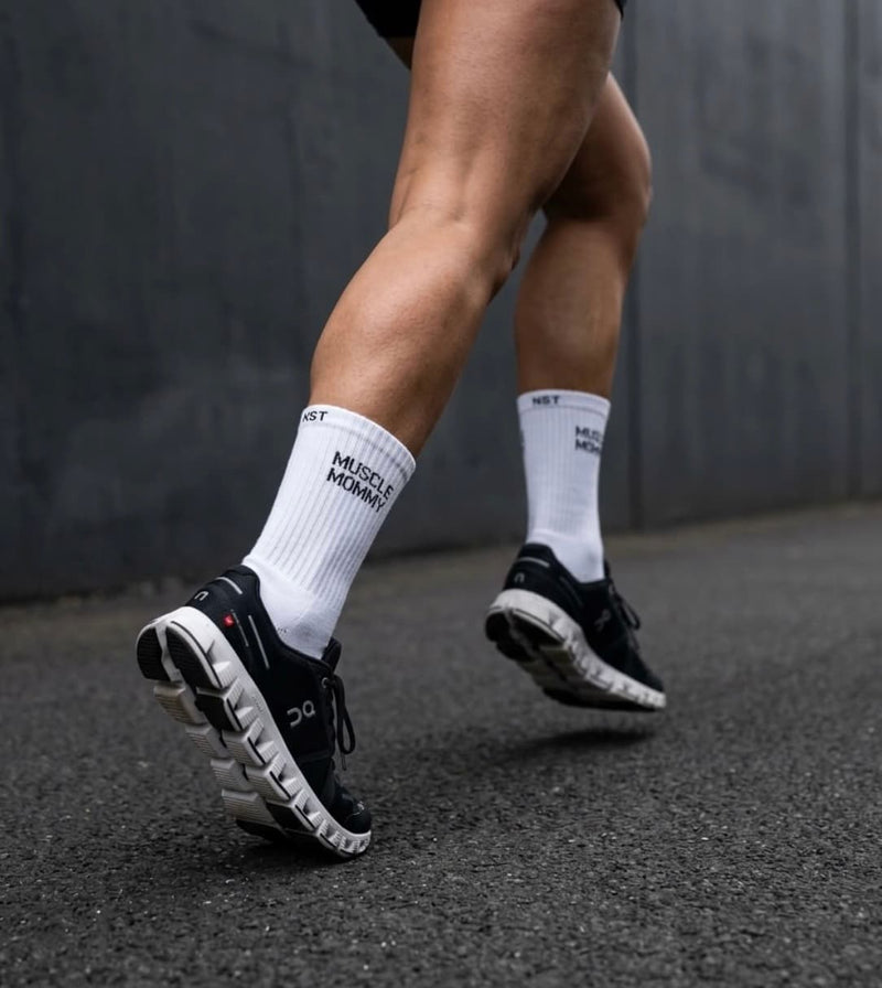 Weiße Sportsocken mit „Muscle Mommy“-Schriftzug beim Laufen in schwarzen Sneakers