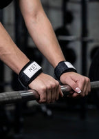Frau hält eine Langhantel mit schwarzen NST Fitness Zughilfen mit grauem Wabenmuster und Logo an beiden Handgelenken. Die Zughilfen unterstützen einen sicheren Griff beim Krafttraining und Gewichtheben.